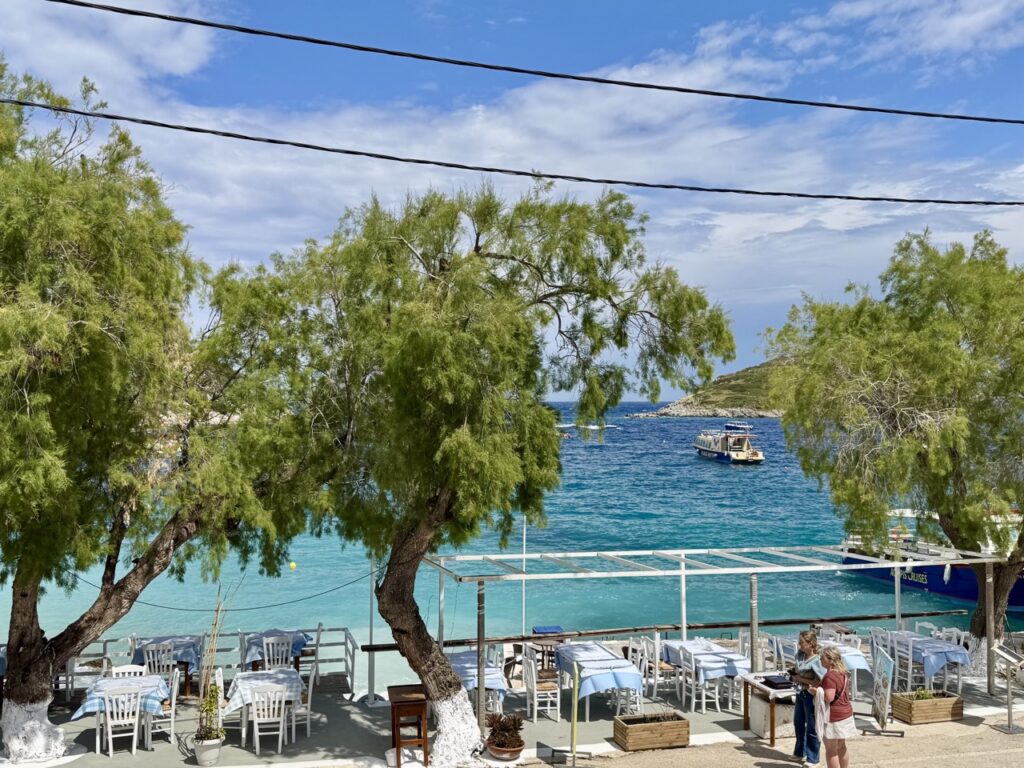 Wakacje 2026 w Grecji. Grecka tawerna z białymi stolikami i widokiem na morze, w tle łódki na błękitnej wodzie