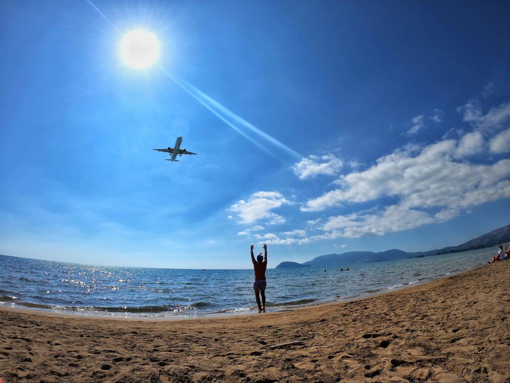 Plaża Kalamaki na Zakynthos z osobą stojącą na piasku i samolotem przelatującym nisko nad morzem