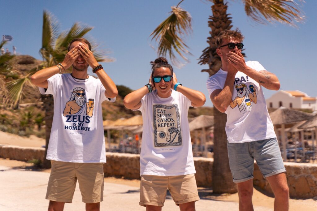 Trzy osoby w greckich koszulkach Magic Tours – Zeus, Gyros Repeat i OPA – na plaży.