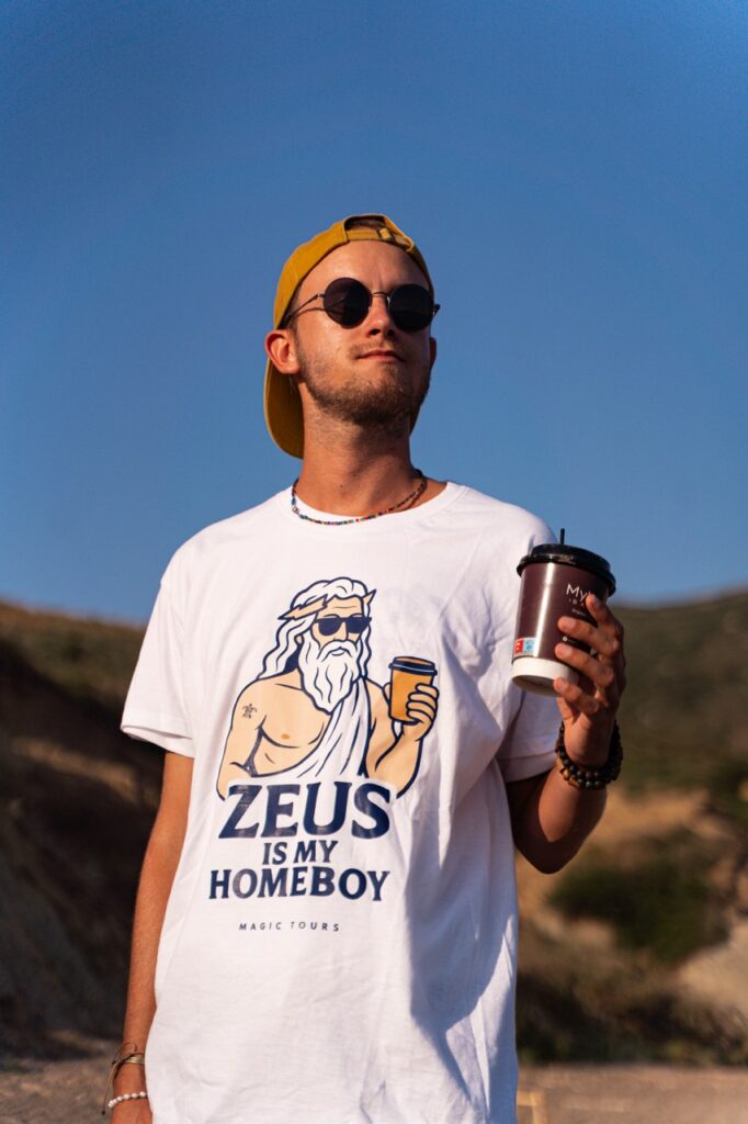 Mężczyzna w koszulce Zeus Is My Homeboy Magic Tours trzymający kawę.