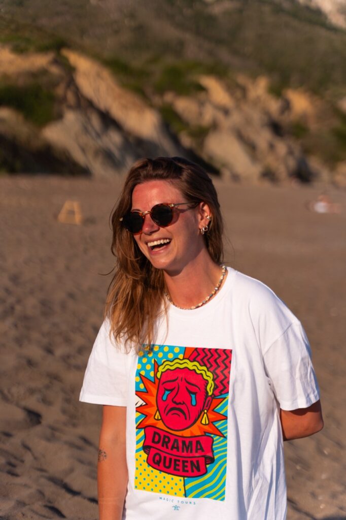 Kobieta w kolorowej koszulce Drama Queen Magic Tours na greckiej plaży.